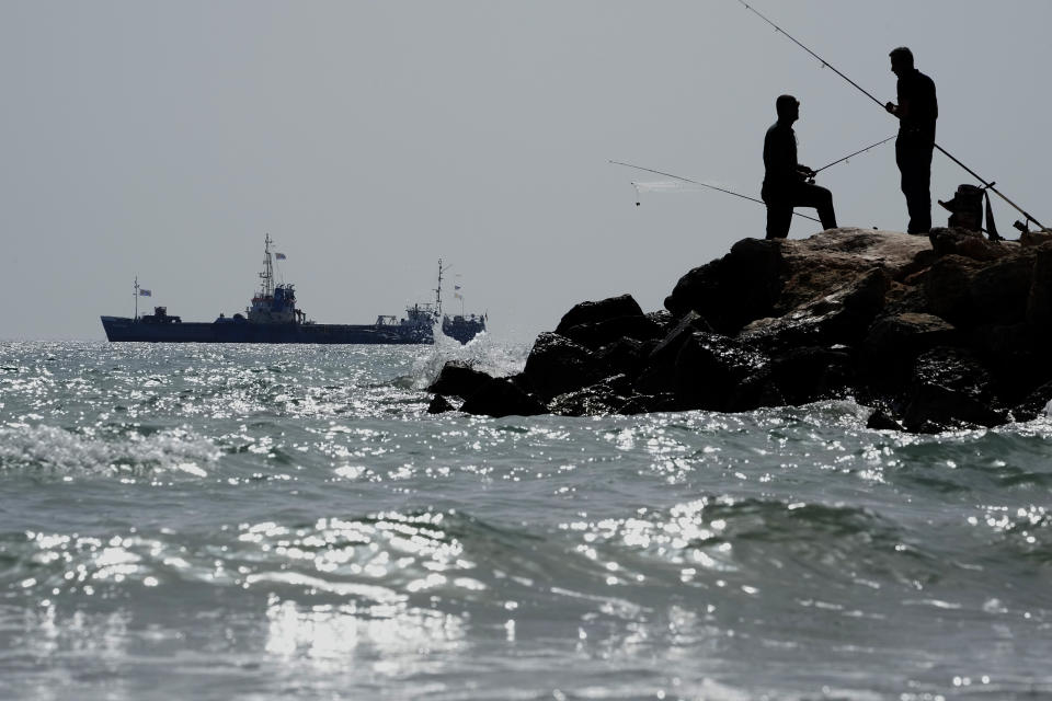 Men fish as in the background is seen a ship loaded with 240 tons of canned food destined for Gaza rests in waters just outside the Cypriot port of Larnaca, Cyprus, on Saturday, March 30, 2024. U.S. charity World Central Kitchen says the ship, named Jennifer, was due to depart following the inaugural voyage of the Cyprus-Gaza sea route earlier this month by the Open Arms vessel that delivered 200 tons of food and water. (AP Photo/Petros Karadjias)