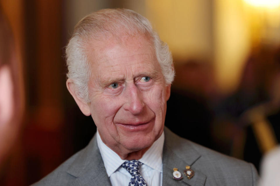 I don't know who this is. An older man in a suit with a patterned tie and lapel pins smiles and looks slightly to the side at an indoor event