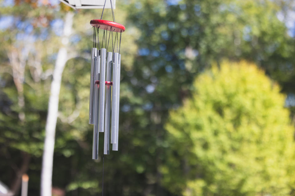 Wind chimes hanging outdoors with a background of lush, green trees. The chimes consist of several metal tubes