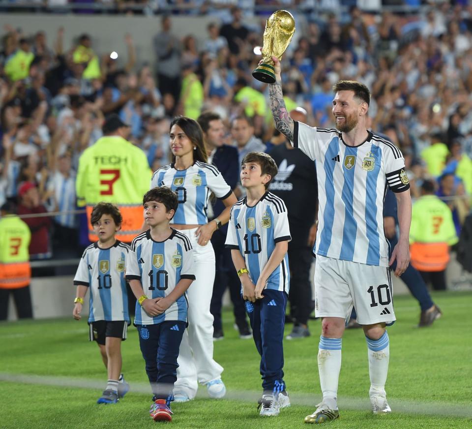 lionel messi, his wife, and three youth sons walk on a soccer field, all are wearing argentina national team jerseys and messi is holding up the fifa world cup trophy, behind them is a crowd in the stands