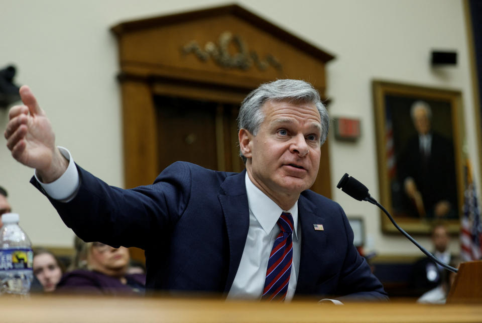 Christopher Wray, at a microphone, holds his right arm out to his side in an apparent gesture.