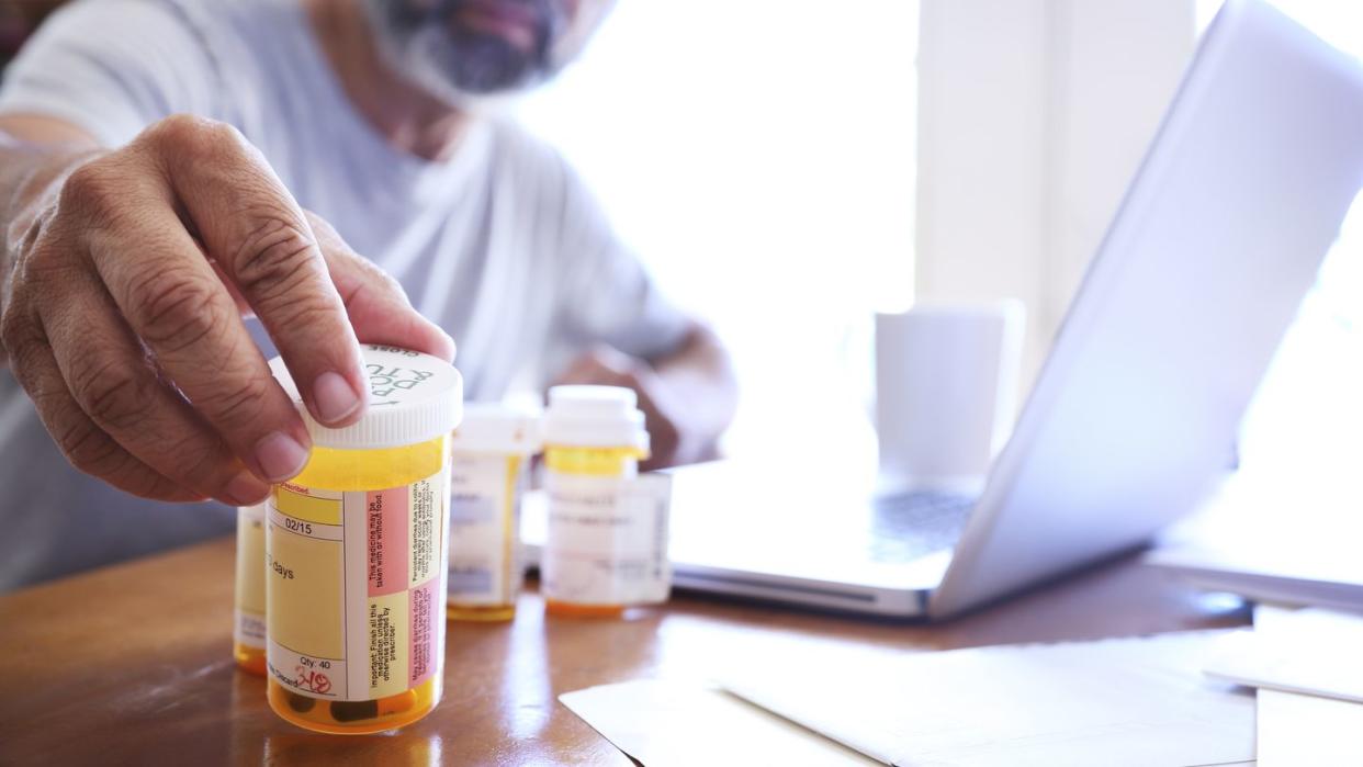 Hispanic Man Sitting At Dining Room Table Reaches For His Prescription Medications