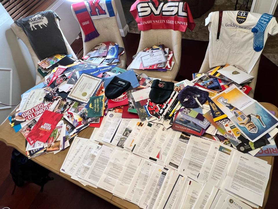 A neatly stacked array of scholarship offer letters and college brochures on a coffee table, with four chairs behind it draped with college swag.