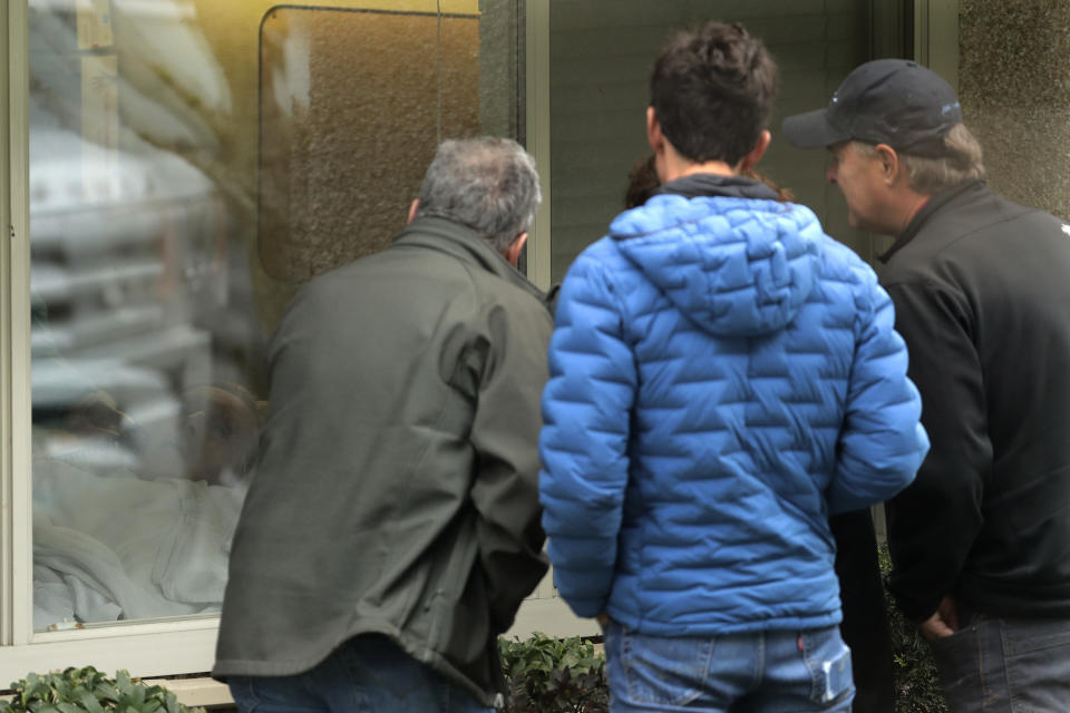 In this March 12, 2020, photo, family members of Chuck Sedlacek, left, look at him through reflections in the window of his room at Life Care Center in Kirkland, Wash., near Seattle. The facility has been at the center of the COVID-19 coronavirus outbreak in the state, and Sedlacek's son Scott said that he and his siblings have barely spoken to their father inside the center, who in addition to testing positive for the coronavirus, has blindness, neuropathy, and has difficulty using a phone, saying he is more of an "inmate" than a patient. Residents of assisted living facilities and their loved ones are facing a grim situation as the coronavirus spreads across the country, placing elderly people especially at risk. (AP Photo/Ted S. Warren)