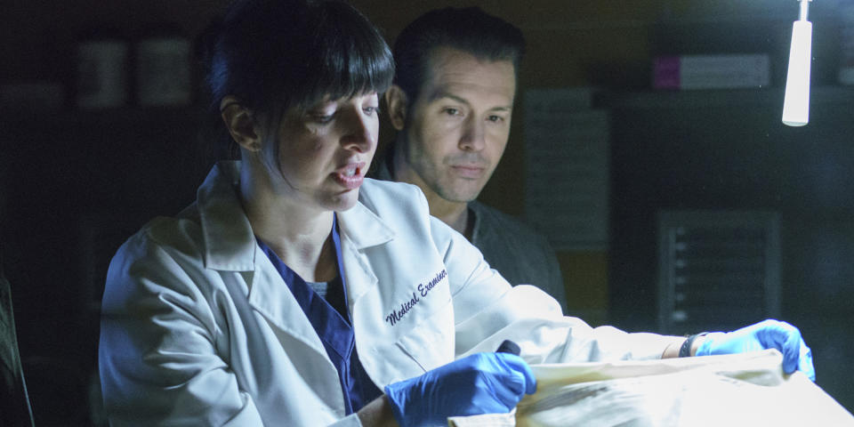 A woman with dark brown hair and bangs looks at a cadaver in a white lab coat that says medical examiner. She's wearing blue medical rubber gloves. (Parrish Lewis / NBCUniversal)