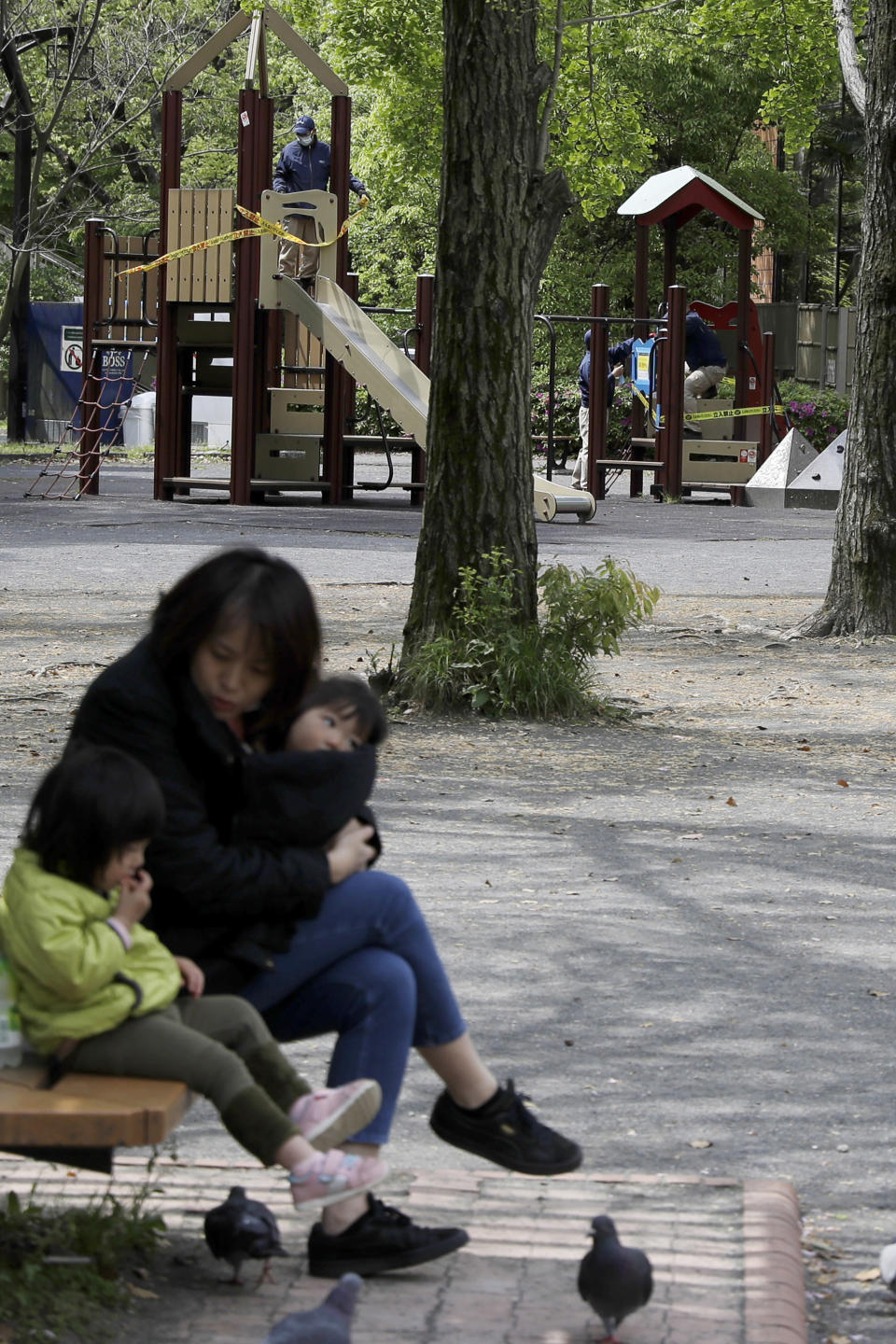 In this April 24, 2020, photo, a woman sits with her children as workers in background apply ribbon reading, do not enter, sealing off a playground in Tokyo. Under Japan's coronavirus state of emergency, people have been asked to stay home. Many are not. Some still have to commute to their jobs despite risks of infection, while others are dining out, picnicking in parks and crowding into grocery stores with scant regard for social distancing. (AP Photo/Gregorio Borgia)