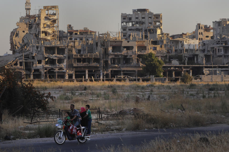 Personas pasan en motocicleta frente a edificios dañados en el pueblo de Homs, Siria, el miércoles 15 de agosto de 2018. (AP Photo/Sergei Grits)
