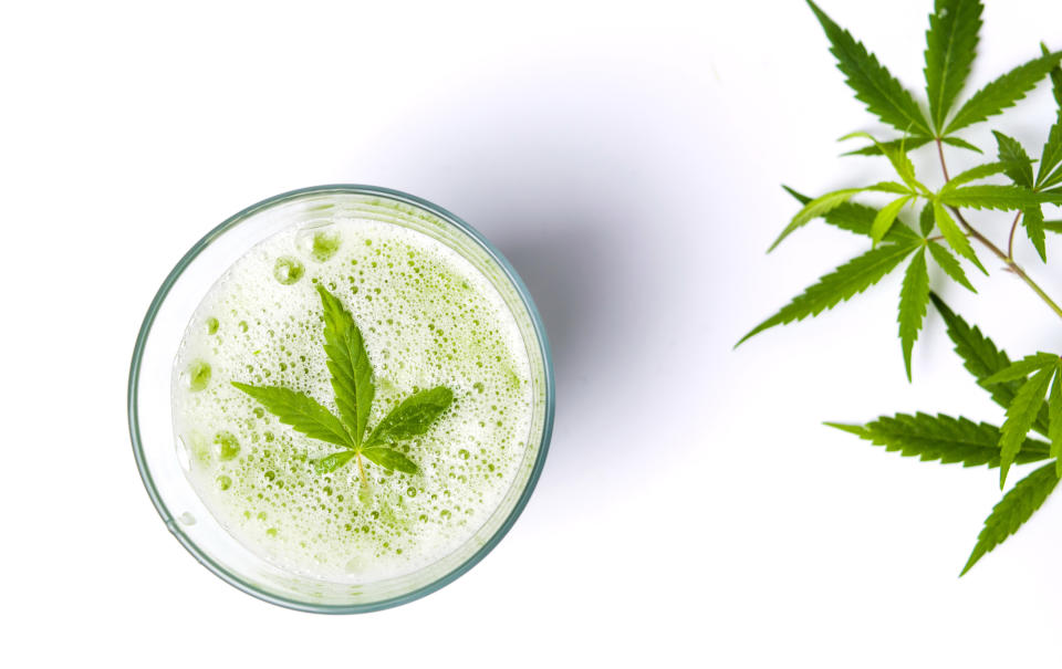 A cannabis leaf lying atop carbonation in a glass, with cannabis leaves to the right of the glass.