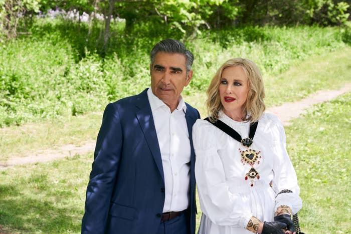 Two individuals stand outdoors, one in a suit and the other dressed in a white outfit with a prominent heart design