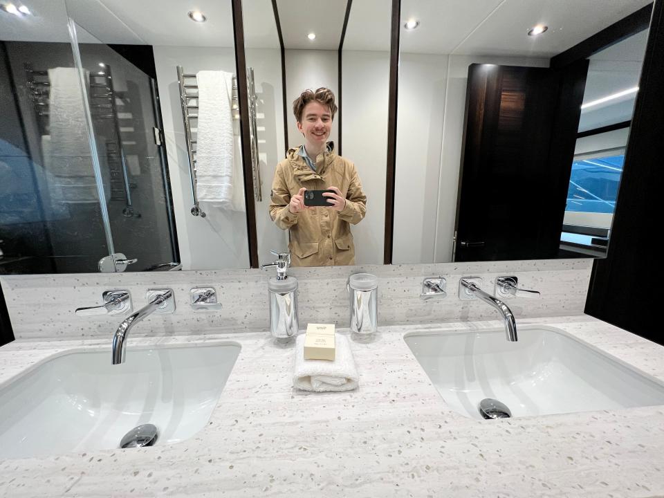 A man takes a selfie in the bathroom onboard a Sunseeker 76,  where white marble countertops decorate two sinks