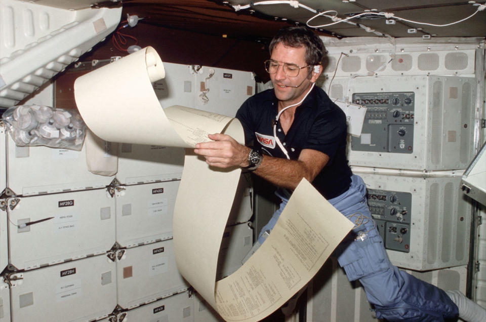 a man in a blue shirt holds a large roll of teleprinter copies while floating aboard a spacecraft