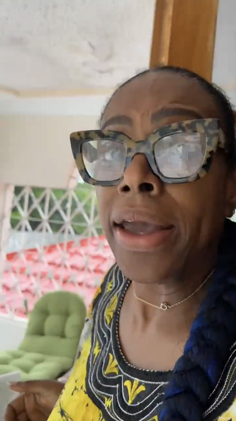 A woman with large glasses and braided hair wears a patterned top and gold necklace, speaking indoors with red and white patterned walls in the background