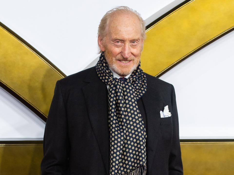 charles dance on the red carpet for the king's man, wearing a black suit coat, black and gold fringed scarf, and white pocket chief