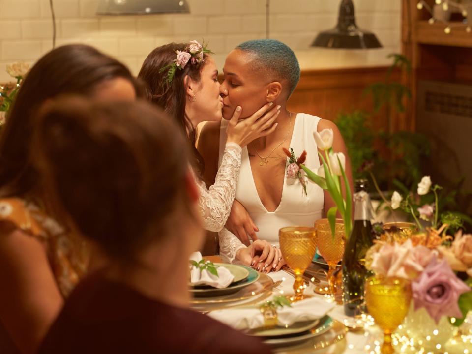 Two brides kiss at a table surrounded by their friends.