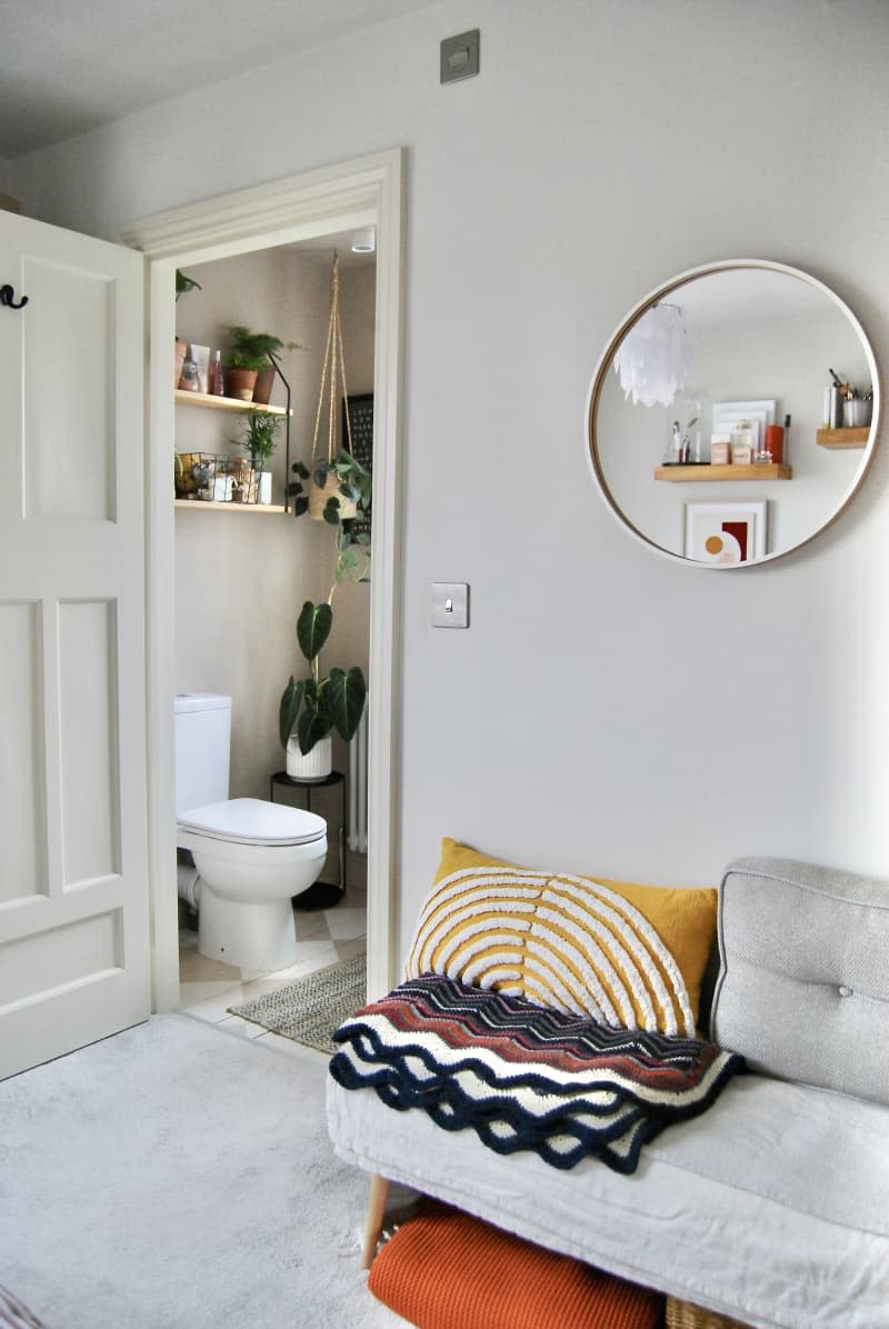 Grey couch with decorative pillows and throw next to bathroom entryway near circular mirror.