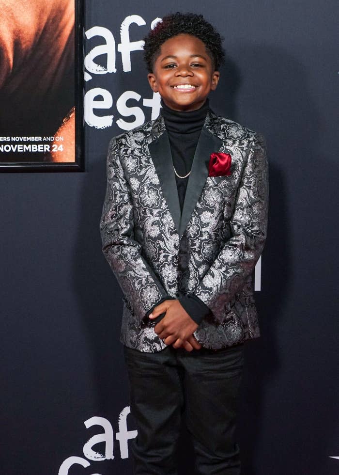 A young boy smiles on a movie premiere carpet with a suit on.