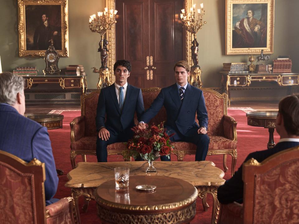 alex and henry in red white and royal blue, holding hands and sitting on an ornate chaise in a lavish, royal room. the king and henry's brother sit in front of them