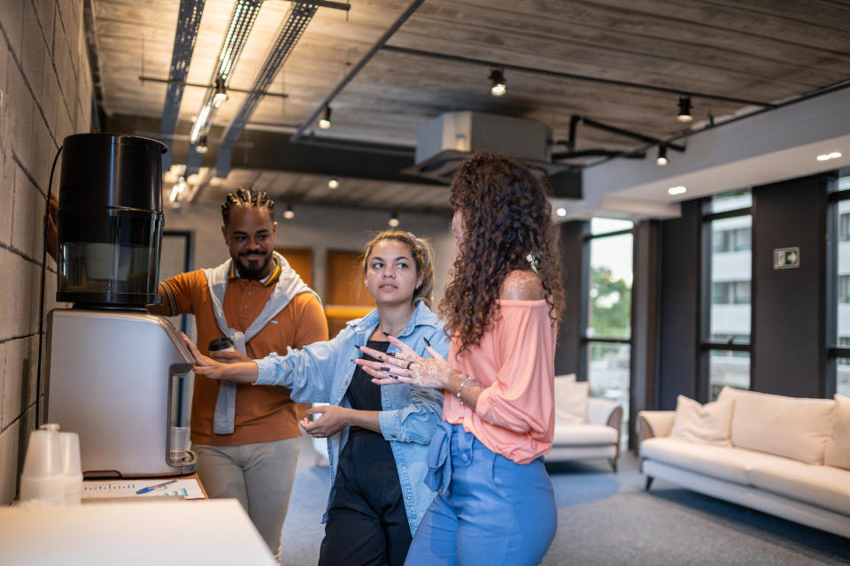 Three colleagues, one man and two women, discussing by a coffee machine in an office lounge area. One woman is gesturing with her hands