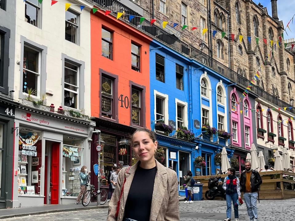 Dasha stands in front of a row of colorful buildings in Edinburgh. Behind her, people are walking around near the shops.