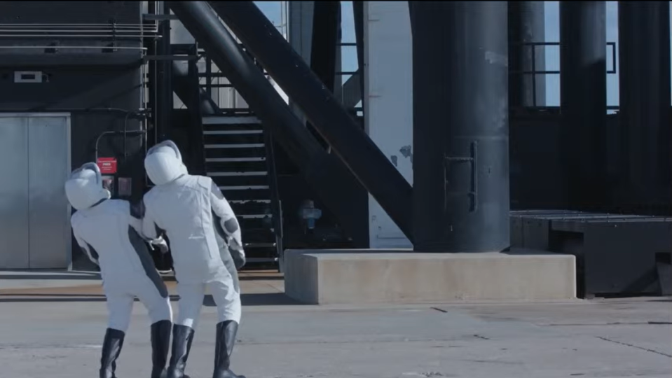 two people in white spacesuits craning their necks back beside a launch pad