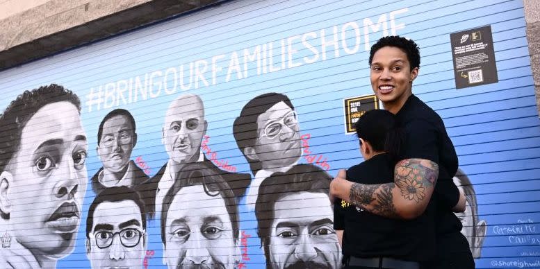 brittney griner hugs someone on her left and looks over her shoulder, she is smiling and both people wear all black outfits, they stand next to a painted mural of 15 people and the slogan bring our families home