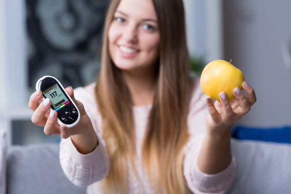 Woman with handheld glucose monitor in one hand and an apple in the other hand.