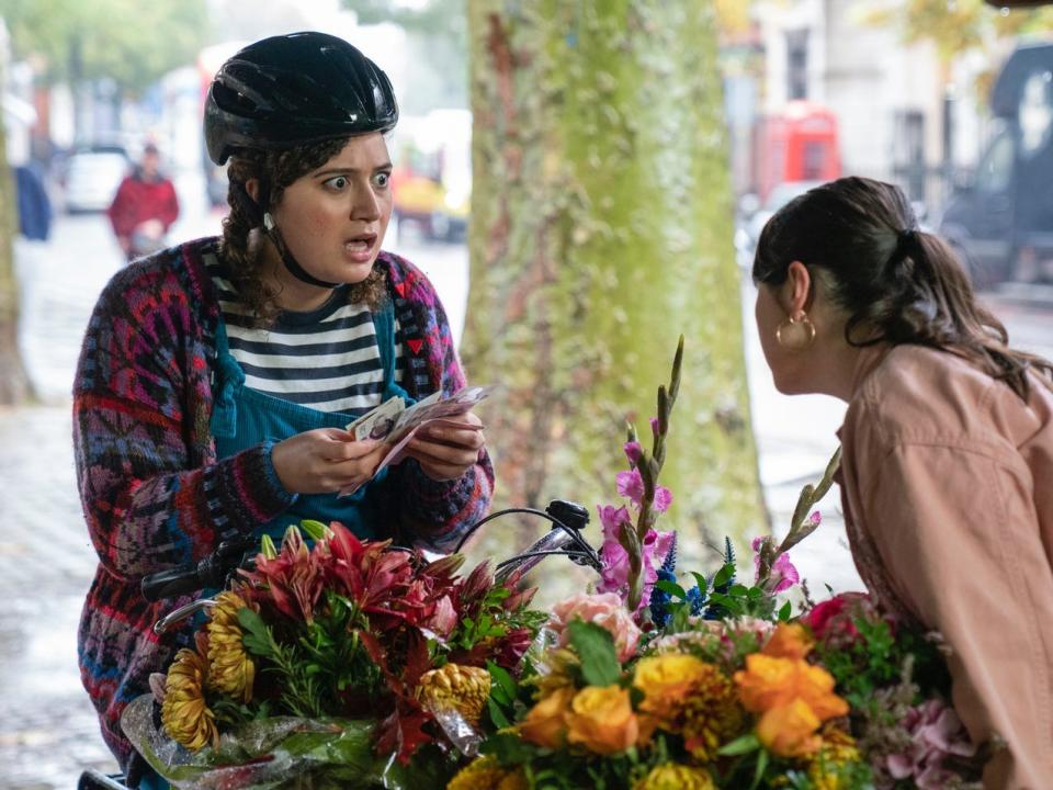 A woman wearing a bike helmet looks shocked as she holds money while speaking to a woman at a flower stand.
