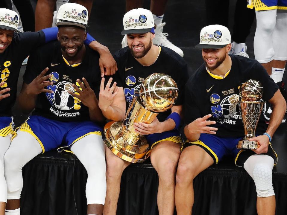 Andre Iguodala, Draymond Green, Stephen Curry, and Klay Thompson sit on a stage next to each other and hold up four fingers.