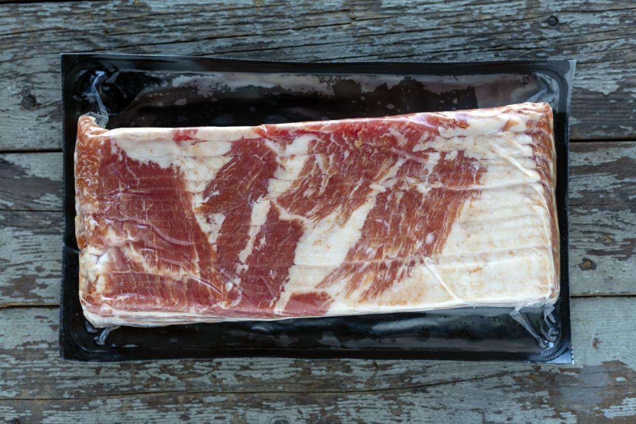 One kilogram of thick sliced smoked bacon wrapped in vacuum sealed plastic packaging on a rustic wood background.