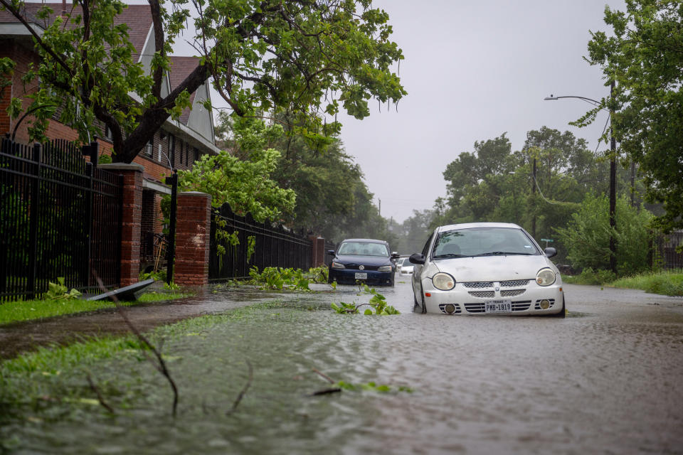 A flooded street with partially submerged cars and debris. Tree branches and leaves are scattered, and the surrounding houses appear affected by heavy rain