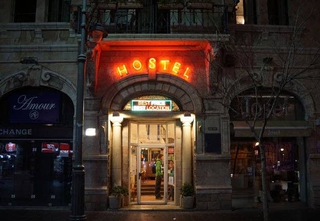 Entrance to a hostel at night with illuminated signs reading "HOSTEL" and "BEST LOCATION." The building has a stone façade, and the street is empty