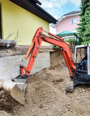 An excavator digs a hole in the ground next to a yellow house. 