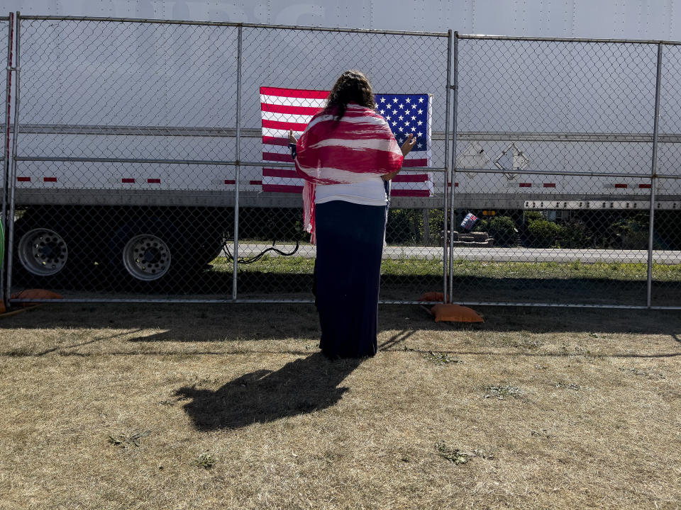 A woman prays as she faces a U.S. flag on the perimeter fence at the ReAwaken America Tour at Cornerstone Church in Batavia, N.Y., Saturday, Aug. 13, 2022. Anthea Butler, a scholar of American religion and politics at the University of Pennsylvania says that that the way Michael Flynn and ReAwaken join Christian nationalism to the idea of spiritual warfare is dangerous because it suggests there are “demonic” people in government, and Christians need to act to save the country. “If people are talking about spiritual warfare and are talking about taking up arms and stuff, then I think you should be very worried,” Butler says. (AP Photo/Carolyn Kaster)