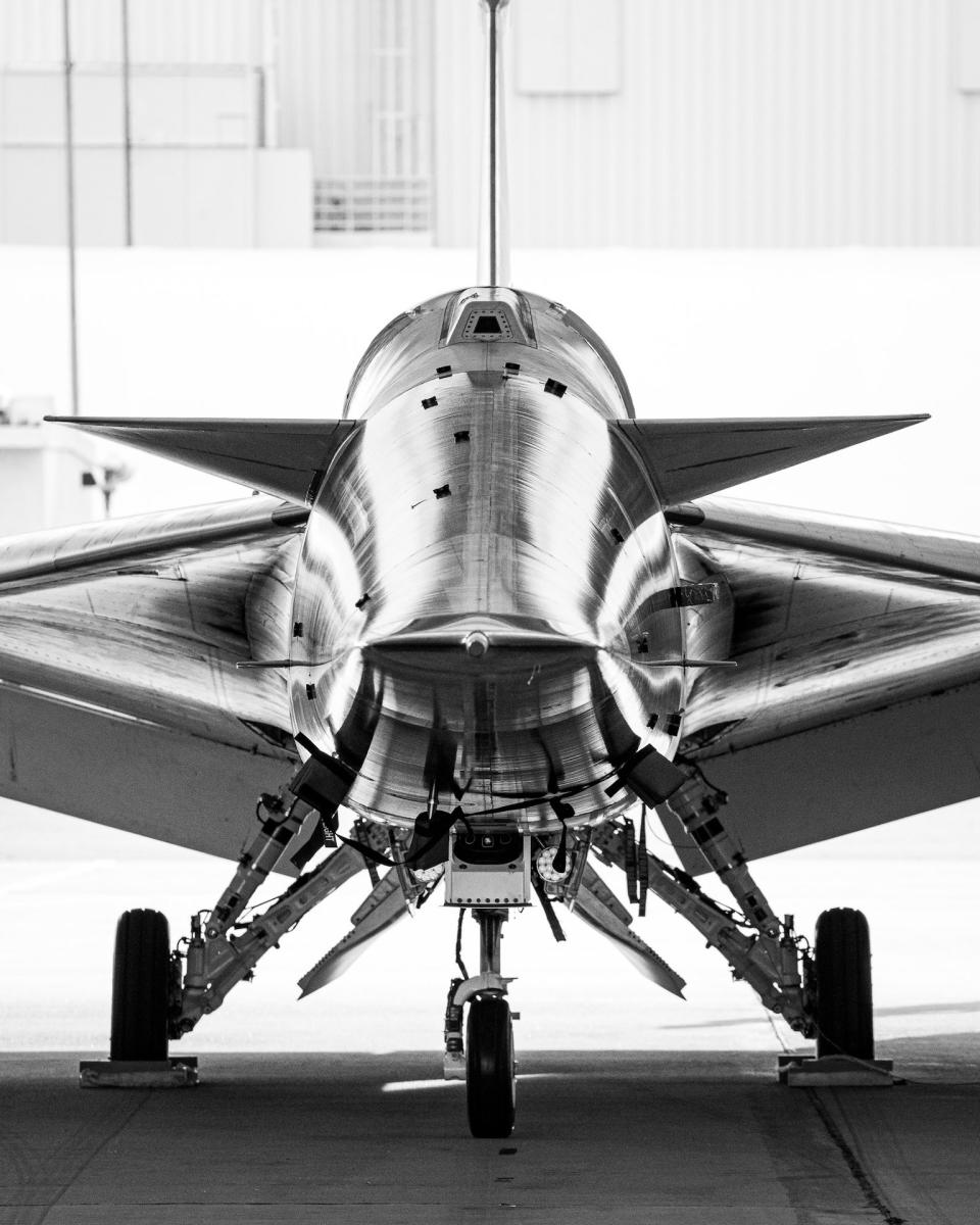 A head-on black and white image of the X-59 supersonic jet. The jet sits on three-wheeled landing gear, and the sharp, sleek angles of the jet's body and wings reflect a s shapely light, highlighting the craft's curves and details.