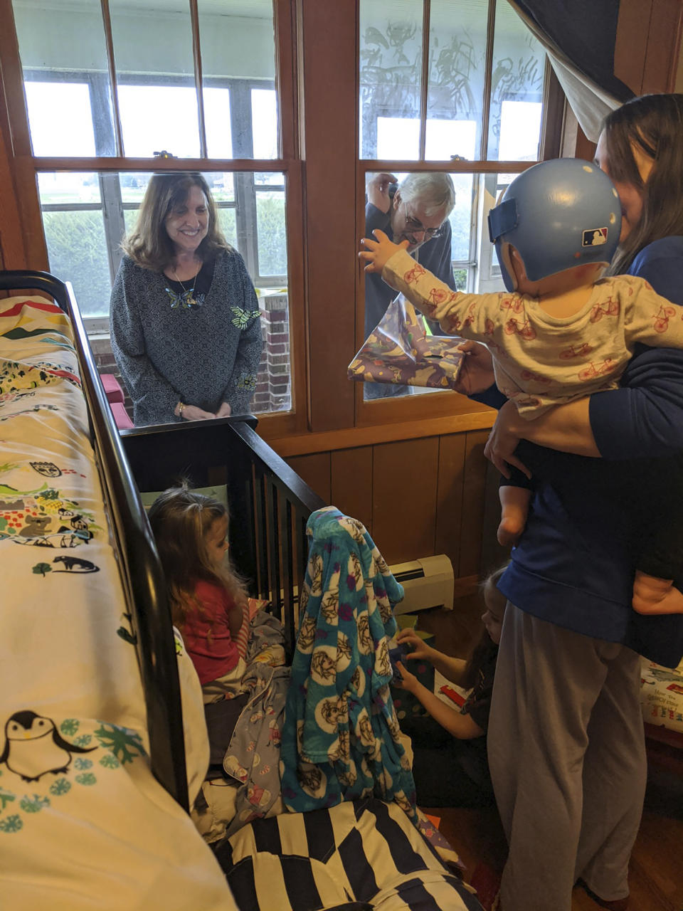 In this April 3, 2020 photo provided by Dann and Heather Voelz, Heather Voelz and her three children get a visit from Heather's parents, who watch their grandchildren open Easter baskets through a window in Taylorville, Ill. The children usually visit their grandparents every day but haven't been able to because of COVID-19. The Voelz family submitted the photo to the Illinois State Museum, which is documenting what daily life is like for Illinois families during the coronavirus pandemic. (Dann Voelz via AP)