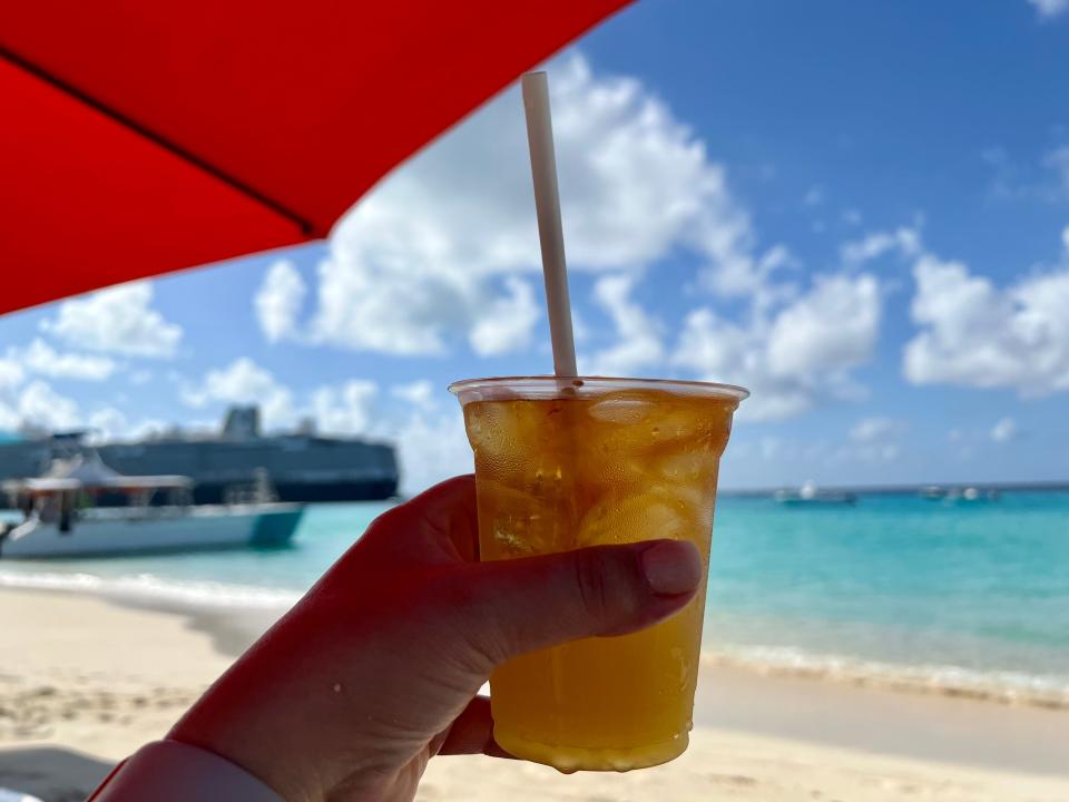 Image of a hand holding a rum cocktail in a plastic cup with a straw. Behind the drink is a beach out of focus and a ship on the left side. Overhead is a red umbrella in the left top corner of the photo.