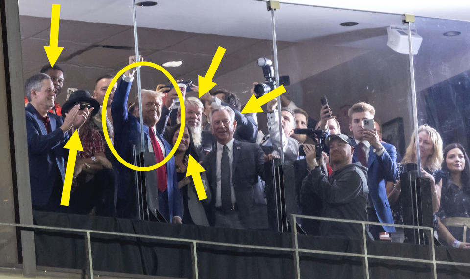 Donald Trump and Melania Trump smile and wave with a crowd of people, some taking photos, behind a glass barrier at an event