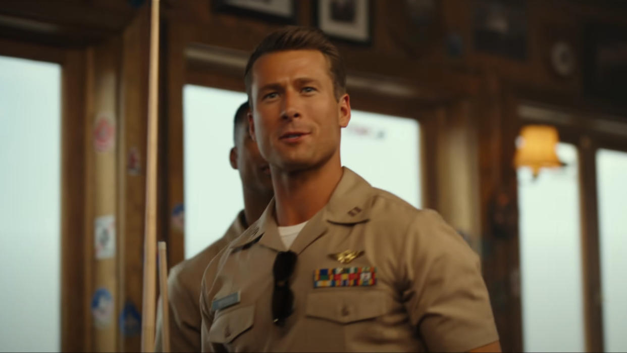  Glen Powell walking and talking with a pool cue in hand in Top Gun: Maverick. 