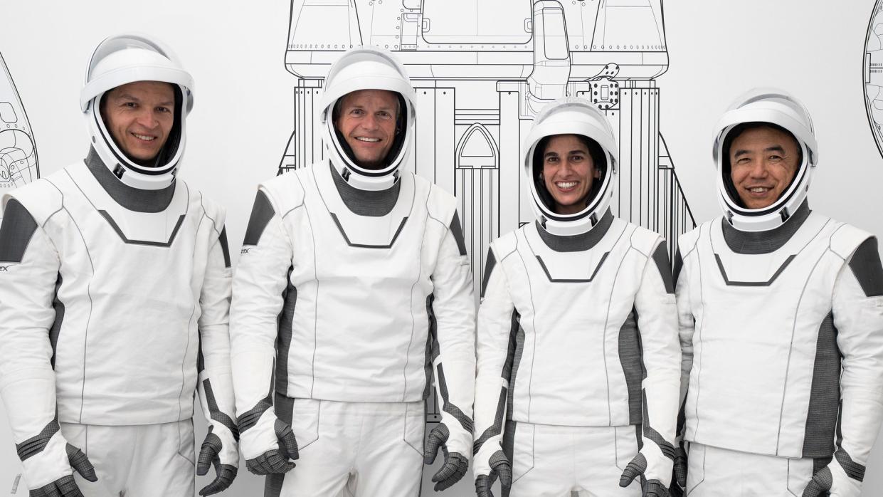  four astronauts standing in a row in white spacesuits and smiling. a line drawing of a rocket is partially visible behind them 