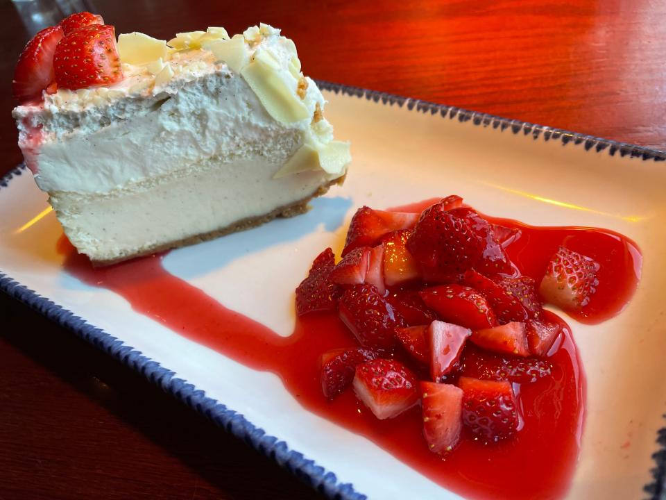 Slice of cheesecake with two layers and white-chocolates flakes with strawberries on top. A pile of strawberries and sauce sits next to the cheesecake on a rectangular plate.