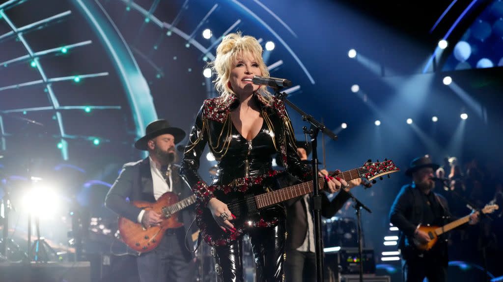 dolly parton smiles while standing behind a microphone on a stand, she plays a black electric guitar and wears a black outfit, two musicians are standing behind her and performing