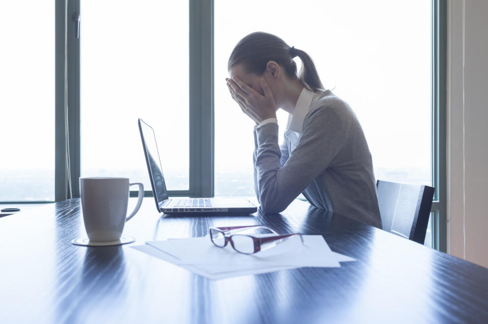 A woman with her head in her hands sits at an office desk with a laptop on it.