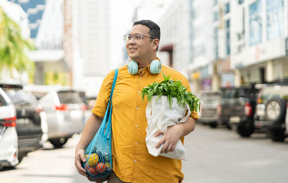 A person wearing glasses and a yellow shirt carries a mesh bag with groceries and a tote bag with leafy greens while walking on a city street