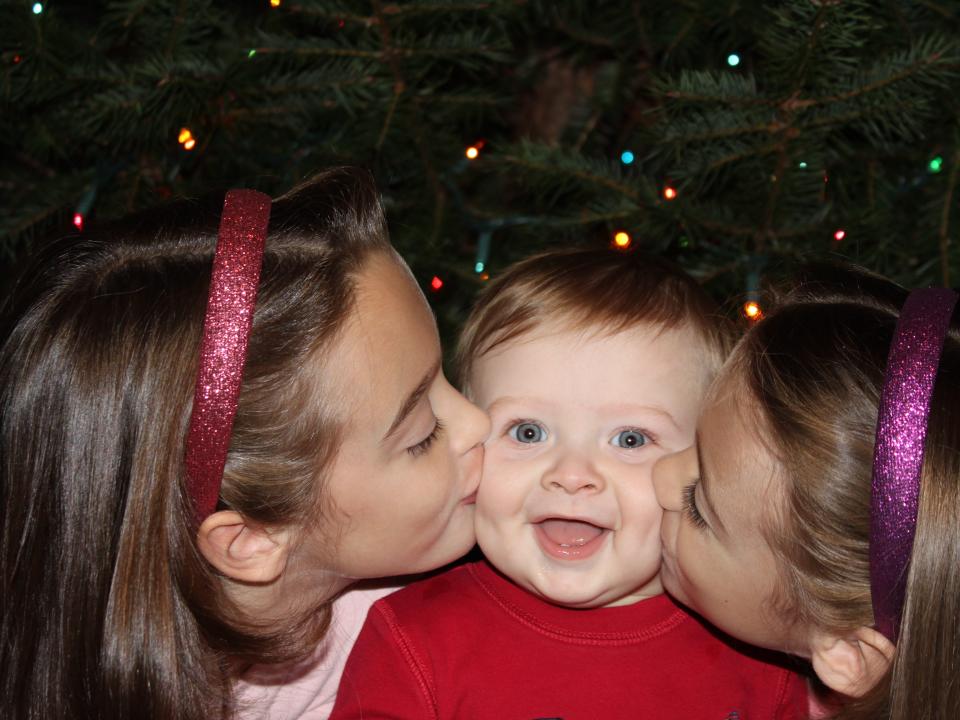 Baby Ben Seitz being kissed by his two older sisters in front of a Christmas tree with multicoloured lights.