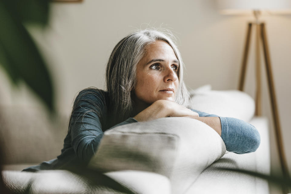 A middle-aged woman with greying hair appears pensive as she rests her chin on her hands while leaning over her sofa 