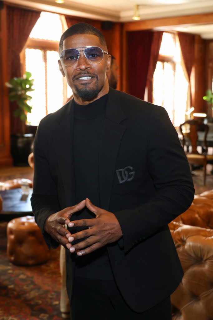 Jamie Foxx stands smiling in a black suit with a logo on the lapel in an elegant room