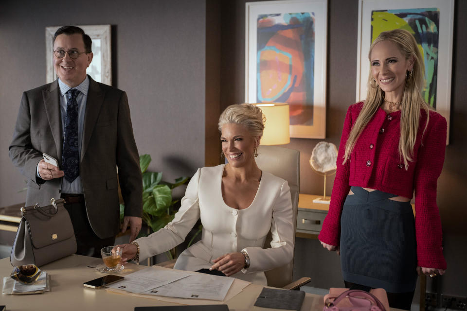 A bespectacled man in a suit and two women in business professional wear smile at someone O.S. in a modern office with abstract wall art, one woman seated and the other two standing; still from "Ted Lasso"