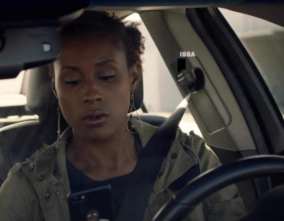 Issa Rae is in a car, wearing a seatbelt and holding a phone while looking at it, with her name shown beside her in the image