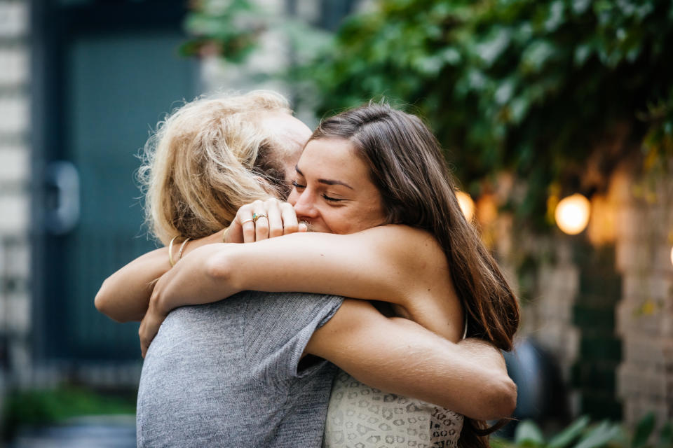 Two people hugging warmly outdoors, one with light hair wearing a gray shirt and the other with dark hair