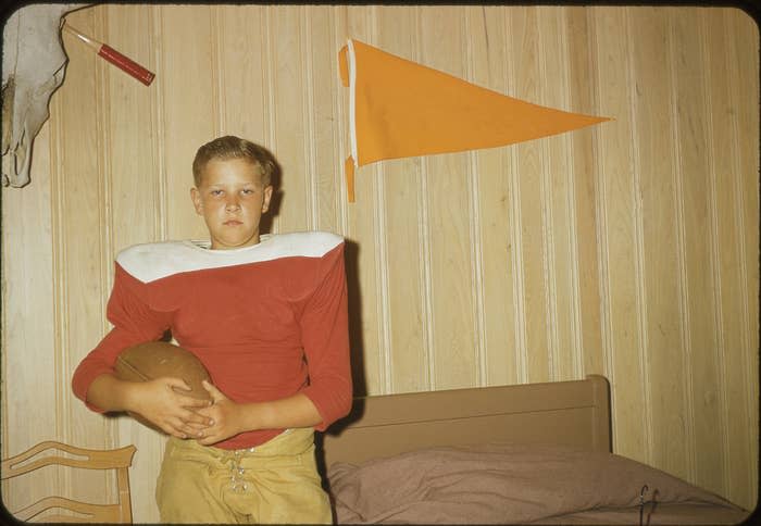 Young boy holds a football while wearing vintage football gear, standing in a wood-paneled room with a pennant on the wall